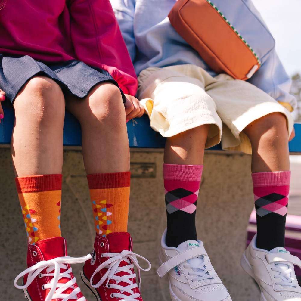 Two children sitting side by side outdoors, showing colorful patterned crew socks paired with sneakers. The child on the left wears orange and red socks with a geometric pattern, and the child on the right wears black, pink, and red socks with an argyle pattern.