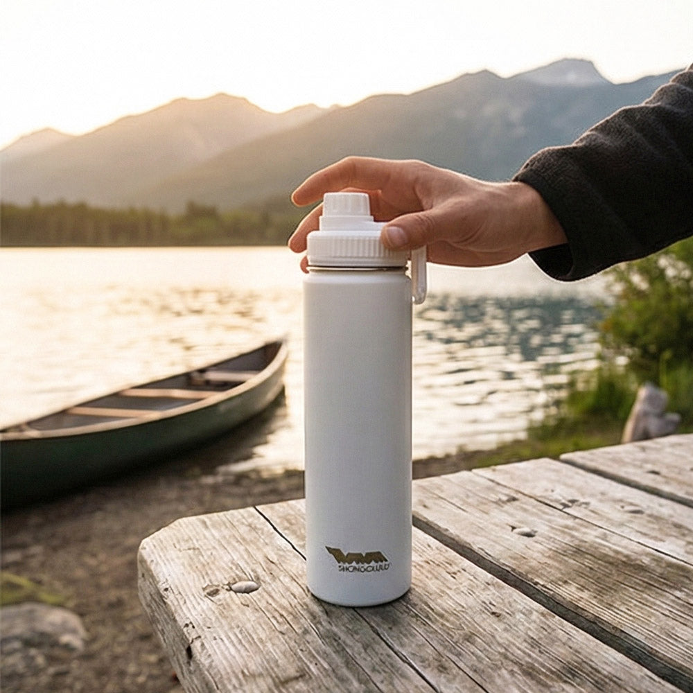 Person holding a white insulated bottle with a scenic lake and mountains in the background