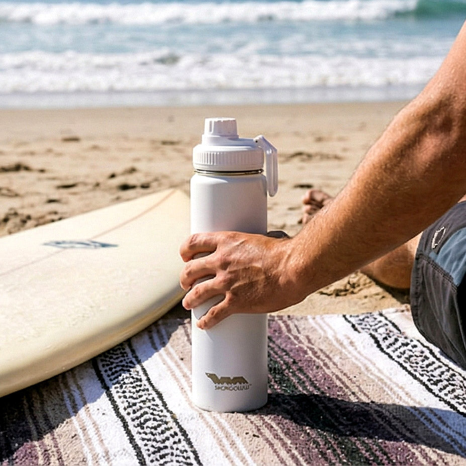 Person holding a white water bottle with a beach and surfboard in the background